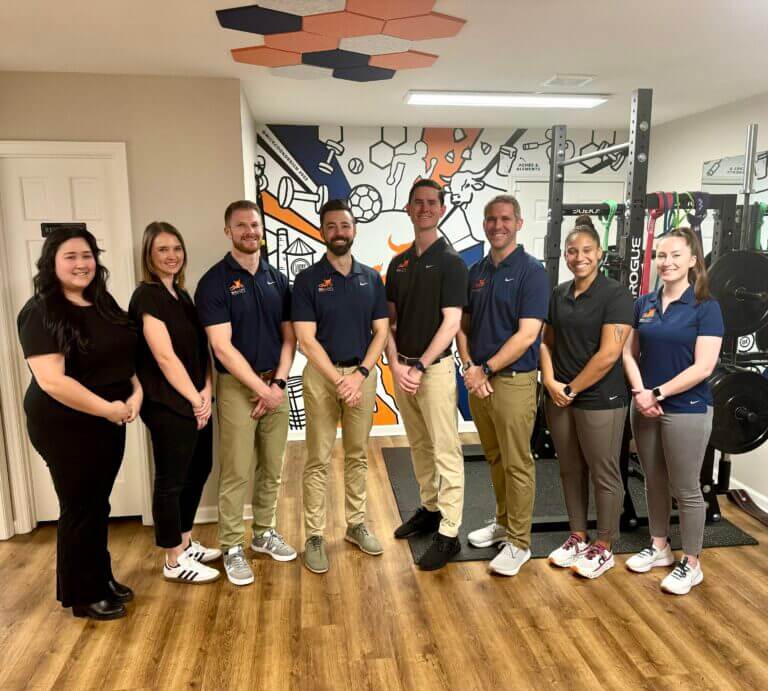 Group of eight fitness trainers smiling in a gym with colorful workout-themed mural and weightlifting equipment in the background.
