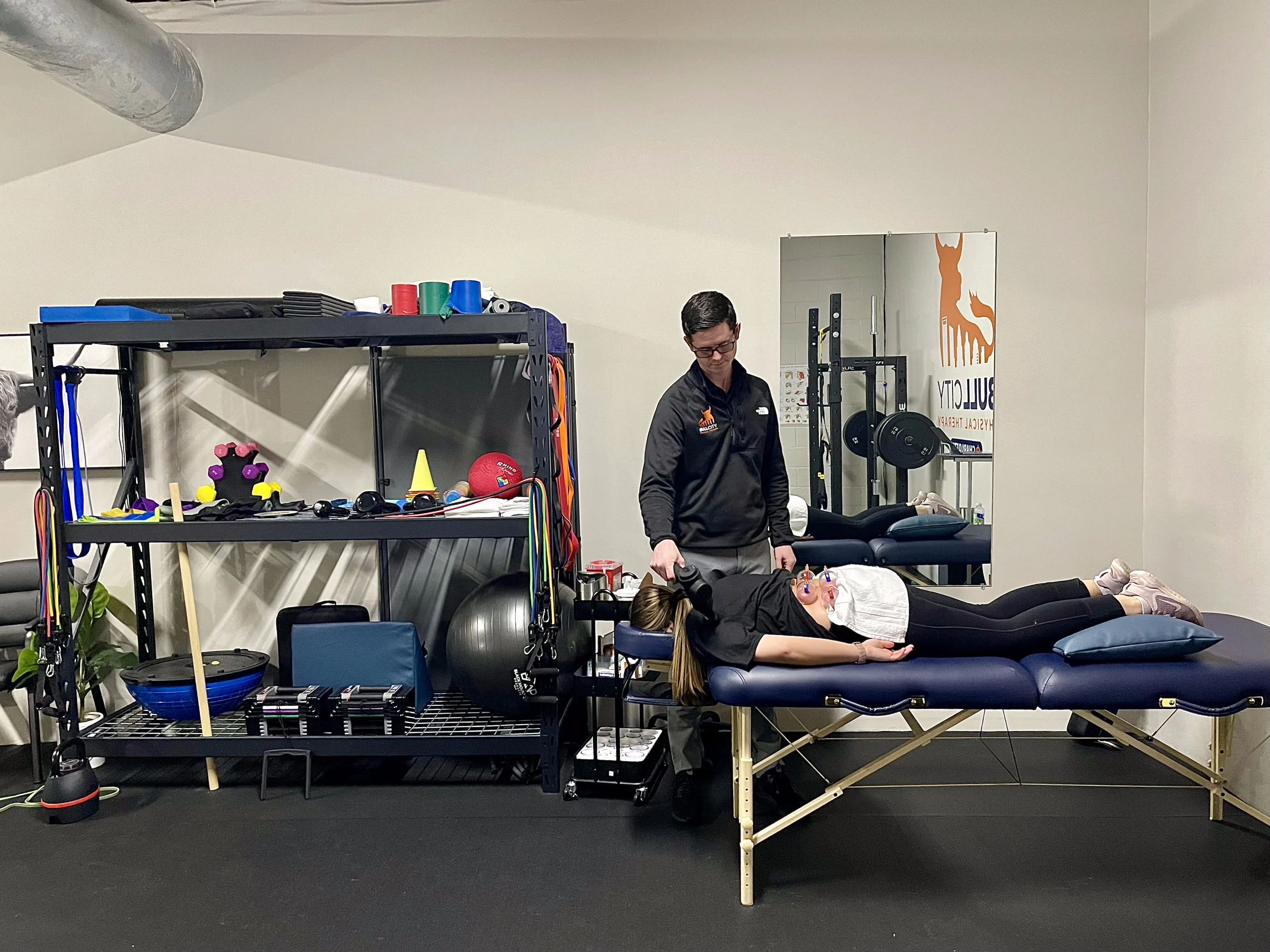 Physical therapist in a clinic adjusting a young woman's arm on a treatment table, surrounded by exercise equipment and rehabilitation tools.
